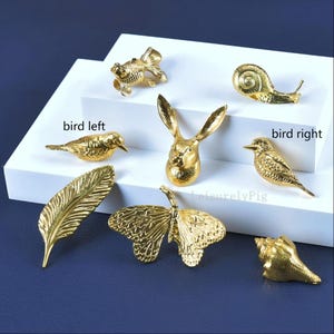 Pu&ograve; includere: Una collezione di figurine decorative color oro, tra cui un pesce, una lumaca, una testa di coniglio, uccelli, una piuma, una farfalla e una conchiglia. Le figurine sono disposte su una superficie bianca su sfondo blu scuro. Il testo "LeisurelyPig" &egrave; visibile.