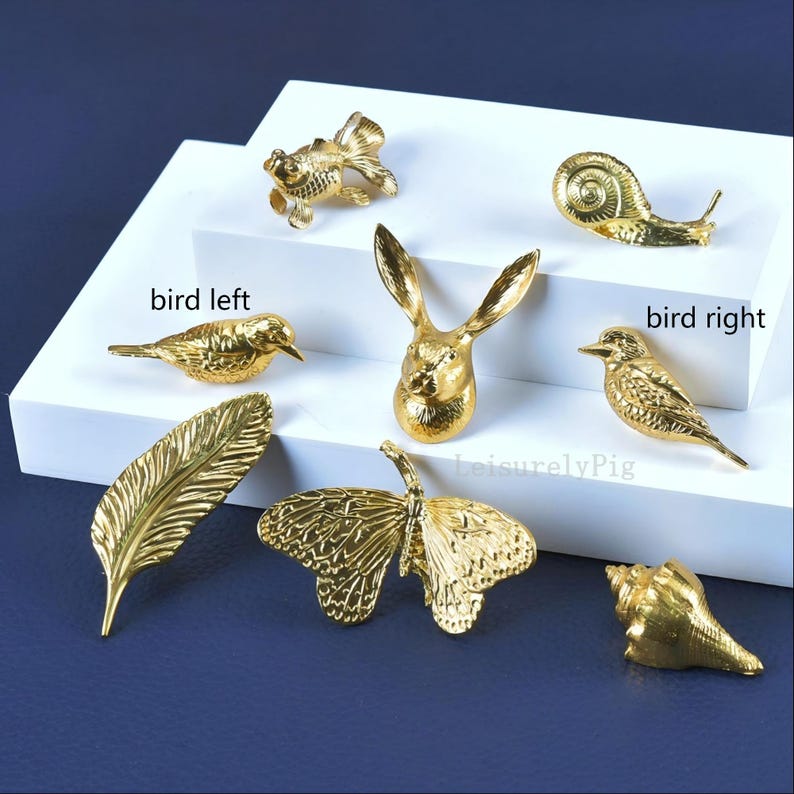 Pu&ograve; includere: Una collezione di figurine decorative color oro, tra cui un pesce, una lumaca, una testa di coniglio, uccelli, una piuma, una farfalla e una conchiglia. Le figurine sono disposte su una superficie bianca su sfondo blu scuro. Il testo "LeisurelyPig" &egrave; visibile.