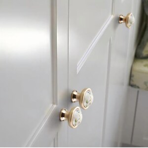 Peut inclure: Gros plan sur une porte d'armoire blanche avec trois boutons de meuble dor&eacute;s. Chaque bouton a un centre en c&eacute;ramique blanche avec un motif floral dans les tons de rose, vert et bleu. Les boutons sont mont&eacute;s sur la porte de l'armoire.