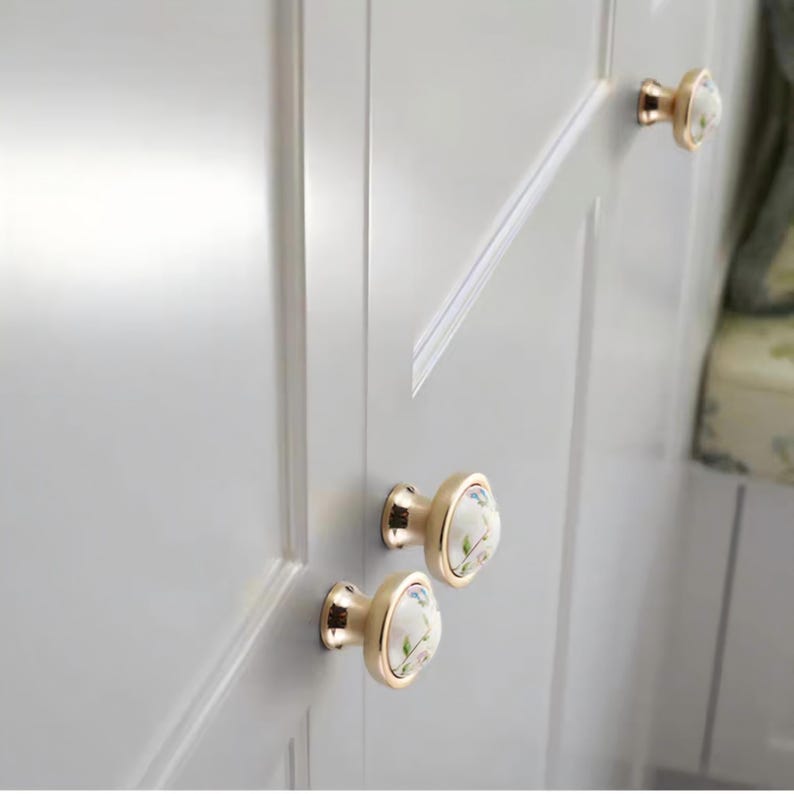 Peut inclure: Gros plan sur une porte d'armoire blanche avec trois boutons de meuble dor&eacute;s. Chaque bouton a un centre en c&eacute;ramique blanche avec un motif floral dans les tons de rose, vert et bleu. Les boutons sont mont&eacute;s sur la porte de l'armoire.