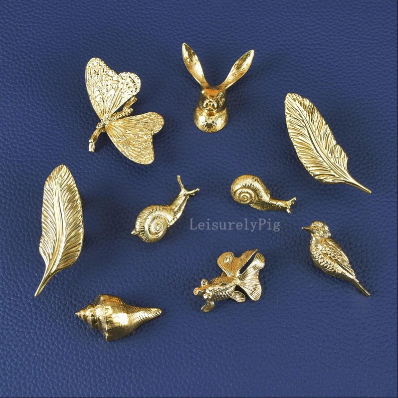 Pu&ograve; includere: Una collezione di piccoli oggetti decorativi color oro, tra cui una farfalla, una testa di coniglio, piume, lumache, una conchiglia, un pesce e un uccello. Gli oggetti sono disposti su una superficie blu. Il testo "LeisurelyPig" &egrave; visibile.