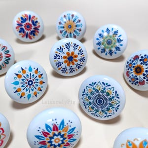 May include: A collection of white ceramic cabinet knobs with colorful floral and geometric designs. The knobs feature various patterns in shades of blue, orange, red, and green, adding a decorative touch to furniture.