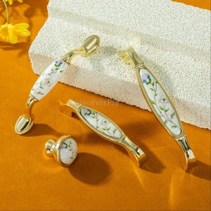 Peut inclure: Ensemble de quincaillerie de meubles dor&eacute;e, comprenant des poign&eacute;es et un bouton. Les inserts en porcelaine blanche sont d&eacute;cor&eacute;s d'un motif floral dans les tons de rose, bleu et vert. Pr&eacute;sent&eacute; sur une surface textur&eacute;e blanc cass&eacute;.