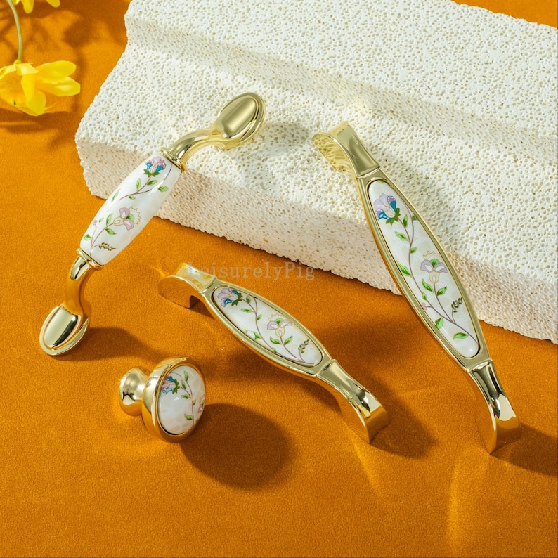Peut inclure: Ensemble de quincaillerie de meubles dor&eacute;e, comprenant des poign&eacute;es et un bouton. Les inserts en porcelaine blanche sont d&eacute;cor&eacute;s d'un motif floral dans les tons de rose, bleu et vert. Pr&eacute;sent&eacute; sur une surface textur&eacute;e blanc cass&eacute;.