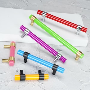 Puede incluir: Una colección de seis tiradores cilíndricos para armarios en varios colores, incluyendo rojo, verde lima, morado, azul, amarillo y rosa. Cada tirador cuenta con una barra de acrílico translúcido sujeta por dos soportes metálicos en acabados dorado, plateado o negro.