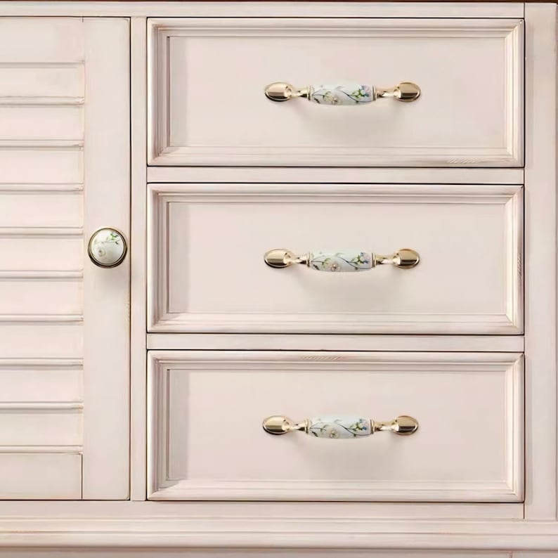 Peut inclure: Un meuble cr&egrave;me avec trois tiroirs et une porte lat&eacute;rale. Les tiroirs ont des poign&eacute;es dor&eacute;es avec des inserts en c&eacute;ramique florale. La porte de l'armoire a un bouton rond en c&eacute;ramique avec un motif floral. Le meuble est peint dans un style vieilli.