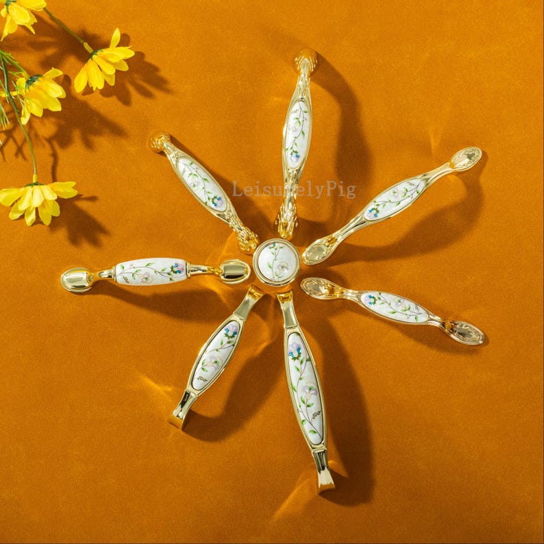 Peut inclure: Ensemble de quincaillerie de meubles dor&eacute;e, dispos&eacute;e en cercle. Chaque pi&egrave;ce pr&eacute;sente une surface en c&eacute;ramique blanche orn&eacute;e de motifs floraux bleus et roses. L'ensemble comprend des boutons et des poign&eacute;es, id&eacute;al pour la r&eacute;novation de meubles.