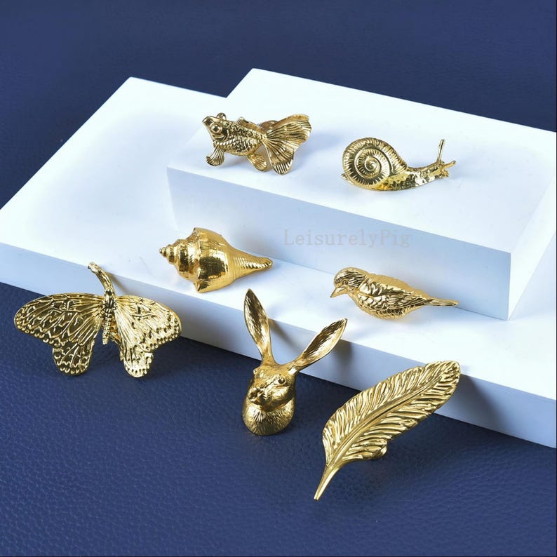 Pu&ograve; includere: Una collezione di figurine decorative color oro, tra cui una farfalla, una lumaca, una conchiglia, un uccello, una testa di coniglio, una piuma e un pesce. Le figurine sono disposte su una superficie bianca su uno sfondo blu scuro.