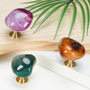 May include: Three cabinet knobs featuring polished, irregular, stone-like tops in purple, amber, and dark green, each mounted on a circular brass-colored metal base. The stones display natural swirling patterns and a smooth, glossy finish.