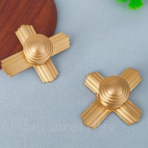 Puede incluir: Dos tiradores dorados para armarios con diseño en forma de cruz. Cada tirador presenta un elemento central redondeado con anillos concéntricos y cuatro brazos radiales con ranuras verticales. Los tiradores están sobre un fondo azul claro.