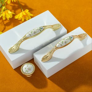 Peut inclure: Ensemble de quincaillerie de meubles dor&eacute;e, comprenant deux poign&eacute;es et un bouton. Les poign&eacute;es ont un design incurv&eacute; avec un motif floral sur fond blanc. Le bouton a un motif floral assorti. La quincaillerie est pr&eacute;sent&eacute;e sur des blocs blancs sur fond orange.