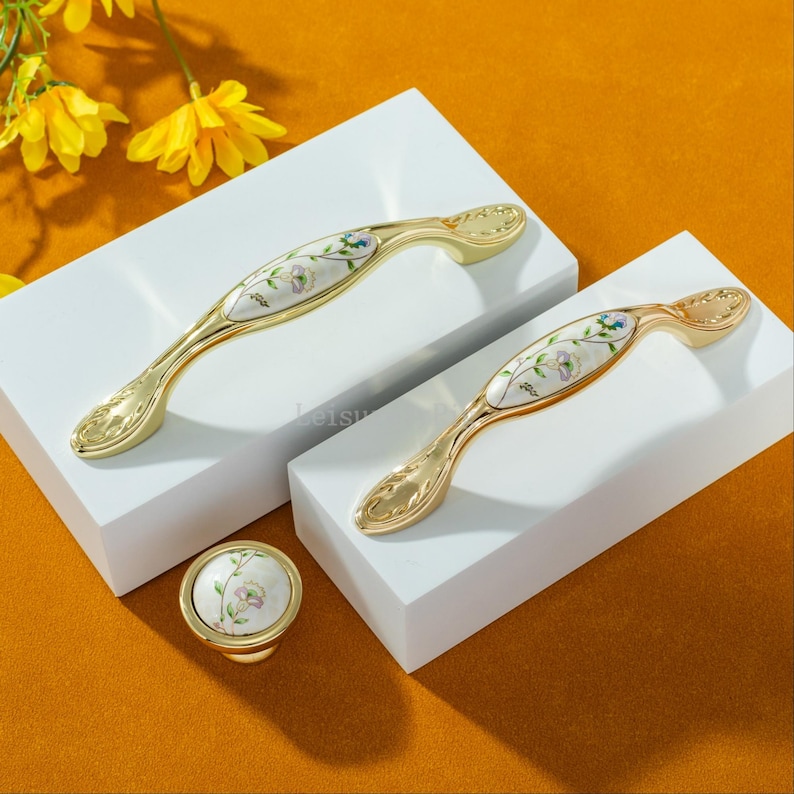 Peut inclure: Ensemble de quincaillerie de meubles dor&eacute;e, comprenant deux poign&eacute;es et un bouton. Les poign&eacute;es ont un design incurv&eacute; avec un motif floral sur fond blanc. Le bouton a un motif floral assorti. La quincaillerie est pr&eacute;sent&eacute;e sur des blocs blancs sur fond orange.