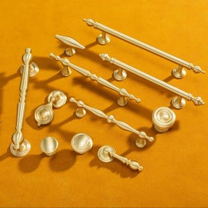 Può includere: Una collezione di maniglie e pomelli dorati per mobili, disposti su una superficie giallo senape. L'hardware presenta vari design, da pomelli semplici a maniglie ornate, adatti per aggiornamenti di mobili, con misure in cm.