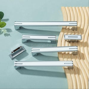 Könnte beinhalten: Eine Sammlung weißer und verchromter Schrankgriffe und -knöpfe. Die Hardware hat ein rechteckiges Design mit einer weißen Mitte und Chromakzenten. Die Griffe variieren in der Größe, mit einem kleinen rechteckigen Knopf und mehreren längeren Griffen. Der Markenname "LeisurelyPig" ist sichtbar.