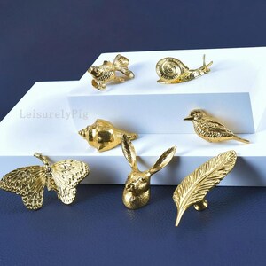 Pu&ograve; includere: Una collezione di pomelli decorativi dorati a forma di pesce, lumaca, conchiglia, uccello, farfalla, coniglio e piuma. I pomelli sono esposti su una superficie bianca su sfondo blu scuro. Il testo "LeisurelyPig" &egrave; visibile.