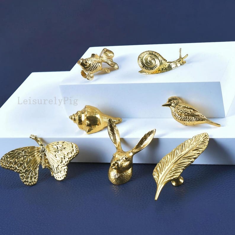 Pu&ograve; includere: Una collezione di pomelli decorativi dorati a forma di pesce, lumaca, conchiglia, uccello, farfalla, coniglio e piuma. I pomelli sono esposti su una superficie bianca su sfondo blu scuro. Il testo "LeisurelyPig" &egrave; visibile.