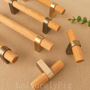 Peut inclure: Poignées de meubles en bois avec des accents métalliques de couleur laiton. Les poignées ont une poignée cylindrique en bois et sont fixées à une base métallique de couleur laiton. Plusieurs poignées sont disposées sur une surface de couleur neutre. Le texte "LeisurelyPig" est visible.