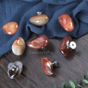 Peut inclure: Plusieurs boutons de meubles en pierre polie, de couleurs rouge, marron et crème. Chaque bouton est doté d'une base en métal argenté. Les boutons sont de formes variées et sont présentés sur une surface en bois foncé.