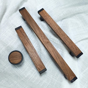 May include: A collection of wooden cabinet handles and a knob. The rectangular handles and the round knob have a dark wood grain finish with black end caps and a black border. The hardware is presented on a white, textured fabric.
