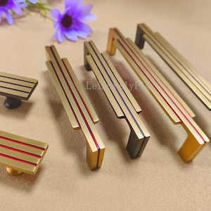 Op de afbeelding: Een verzameling goudkleurige kastbeslagen, waaronder rechthoekige knoppen en stangtrekkers. De hardware heeft rode en blauwe accentstrepen. De knoppen hebben een donkere basis, terwijl de trekkers een verscheidenheid aan donkere en goudkleurige uiteinden hebben.