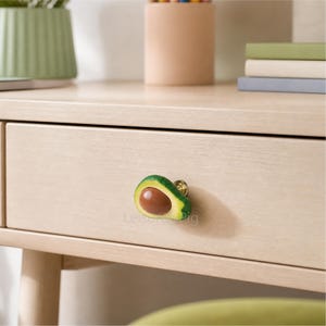 May include: A drawer pull in the shape of a sliced avocado, with a brown pit and green exterior. The drawer pull is attached to a light-colored wooden drawer. The avocado-shaped pull adds a unique touch to furniture.