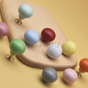 Op de afbeelding: Een verzameling kleurrijke keramische kastknoppen in verschillende tinten, waaronder groen, blauw, rood en geel. Elke knop heeft een ronde vorm en een messing voet. De knoppen zijn gerangschikt op een houten snijplank. Afmetingen in centimeters.