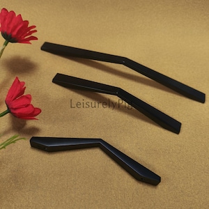 May include: Three black cabinet pulls with an angled design are displayed on a tan surface. The pulls are made of a matte black material. Red flowers are in the background. The text "Leisurely Pig" is visible on the surface.