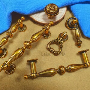 Puede incluir: Una colección de herrajes de latón de estilo antiguo, que incluye tiradores de cajones, pomos y manijas. Los herrajes presentan detalles ornamentados y un acabado dorado cálido. Los artículos están dispuestos sobre una superficie neutra con un paño azul.