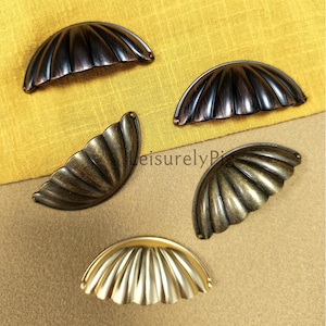 Może przedstawiać: Kolekcja dekoracyjnych uchwytów do szuflad w kształcie muszli w różnych metalicznych wykończeniach. Uchwyty są w odcieniach brązu, antycznego mosiądzu i złota, z żebrowanym wzorem. Prezentowane na żółtym i beżowym tle.