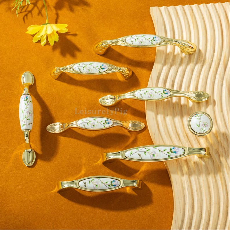 Peut inclure: Ensemble de poign&eacute;es et boutons de placard dor&eacute;s avec des centres en c&eacute;ramique blanche. La c&eacute;ramique pr&eacute;sente un motif floral d&eacute;licat en bleu, vert et violet. La quincaillerie est dispos&eacute;e sur une surface en bois et un fond de couleur moutarde.