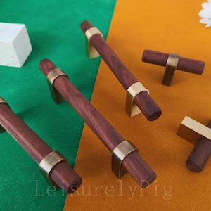 Może przedstawiać: Drewniane uchwyty i gałki do szafek z mosiężnymi akcentami. Uchwyty są w kolorze głębokiego brązu, a gałki mają kształt litery T. Okucia są prezentowane na zielono-pomarańczowym tle. Widoczny jest tekst "LeisurelyPig".