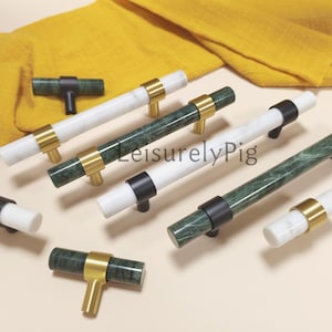 May include: A selection of cabinet handles and knobs in different styles. The handles have white and green marble cylinders with gold, black, or brass details. The knobs are T-shaped with a green marble finish and black hardware.