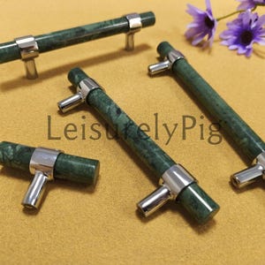 Puede incluir: Un conjunto de tiradores de armario de mármol verde con herrajes plateados. Los tiradores son cilíndricos con un acabado pulido, y los herrajes son de diseño sencillo en forma de T. La imagen incluye el texto "LeisurelyPig".