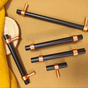 May include: A selection of black cabinet pulls with copper-coloured accents. The pulls have a cylindrical design with copper rings and T-shaped handles. The hardware is arranged on a tan surface with a yellow textile and a wooden slice.