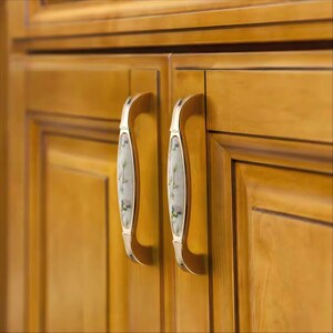 Peut inclure: Gros plan sur deux portes de placard avec des poign&eacute;es d&eacute;coratives. Les poign&eacute;es sont dor&eacute;es avec un centre blanc orn&eacute; d'un motif floral bleu et vert. Les portes sont en bois avec une finition dor&eacute;e et chaleureuse.