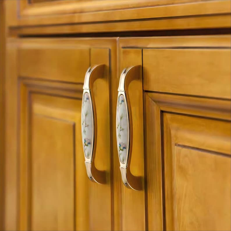 Peut inclure: Gros plan sur deux portes de placard avec des poign&eacute;es d&eacute;coratives. Les poign&eacute;es sont dor&eacute;es avec un centre blanc orn&eacute; d'un motif floral bleu et vert. Les portes sont en bois avec une finition dor&eacute;e et chaleureuse.