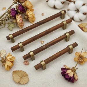 Può includere: Quattro maniglie cilindriche per armadietti in legno marrone scuro con venature visibili, ciascuna dotata di due staffe di montaggio in ottone spazzolato. Le maniglie sono disposte in diagonale, accompagnate da fiori secchi viola e gialli.
