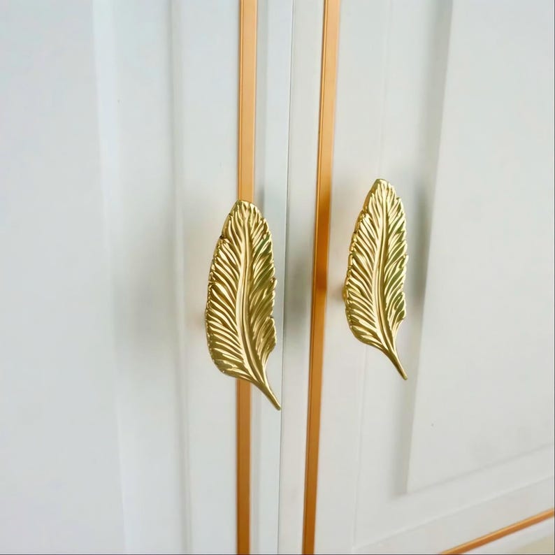 Pu&ograve; includere: Primo piano di un'anta di armadio bianca con due maniglie dorate a forma di piuma. Le maniglie presentano dettagliati motivi a venature e l'armadio ha accenti verticali dorati e bianchi.