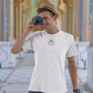 Puede incluir: Una camiseta blanca con un gráfico de cara sonriente negra. La persona que lleva la camiseta está sosteniendo una cámara vintage y sonriendo. La camiseta tiene un tatuaje negro en el brazo que dice "LOVE".