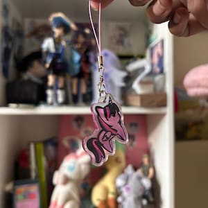 以下が含まれることがあります： 紫とピンクのポニーのキャラクターが描かれた透明なアクリルチャーム。濃い紫色のたてがみと尻尾が特徴です。チャームは銀色の金属製ロブスタークラスプと、吊り下げ用の細い薄ピンク色のコードループに取り付けられており、光沢のある仕上がりになっています。