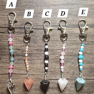 May include: Five metal lobster clasp keychains labeled A through E, each featuring a vertical strand of assorted round beads ending in a pointed, faceted stone pendant. The beads vary in color, including pink, blue, peach, black, white, and clear tones.