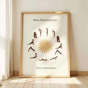 Può includere: Una stampa incorniciata con sfondo beige, un motivo a raggiera dorata e una sequenza di posizioni yoga. La stampa include le parole "Sun Salutation" e "Surya Namaskar" in testo marrone, ideale per gli appassionati di yoga.