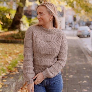 Può includere: Un maglione di lana a treccia marrone con un motivo a rombi. Il maglione ha un collo rotondo e maniche lunghe.