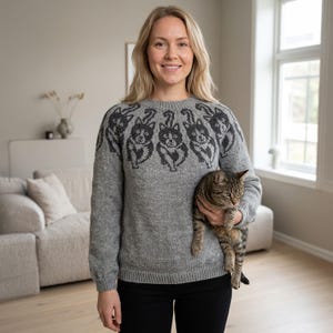 Könnte beinhalten: Ein grauer Pullover mit einem Muster aus schwarzen Katzen auf Brust und Schultern. Der langärmlige Pullover ist aus weichem Strickmaterial. Die Person hält eine getigerte Katze.