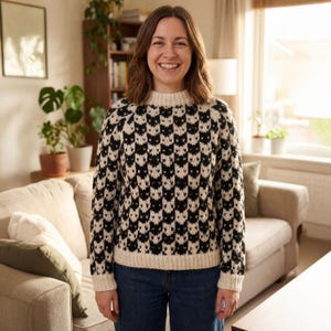 Può includere: Una persona indossa un maglione a fantasia bianca e nera con colletto e polsini color crema. Il maglione presenta un motivo ripetuto di forme nere su sfondo bianco. Indossa jeans blu e sorride.