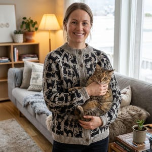 Può includere: Un cardigan grigio con un motivo di gatti neri. Il maglione con bottoni è indossato da una persona che tiene in braccio un gatto tigrato. Il gatto ha il pelo marrone e nero. Lo sfondo include un divano grigio, una lampada e una finestra.