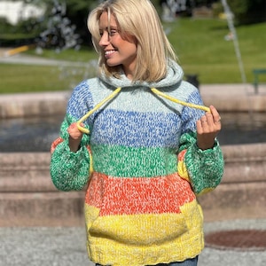 Könnte beinhalten: Ein mehrfarbiger Kapuzenpullover mit Streifenmuster. Der Pullover hat ein hellblaues, blaues, grünes, orangefarbenes und gelbes Design. Er hat eine Kapuze mit Kordelzug und lange Ärmel. Der Pullover ist aus einem dicken, gestrickten Material.