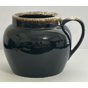 May include: A glossy black ceramic jar with a rounded body and a large handle. The rim and handle have a speckled brown and cream colour. The jar is likely used for storage or decoration.