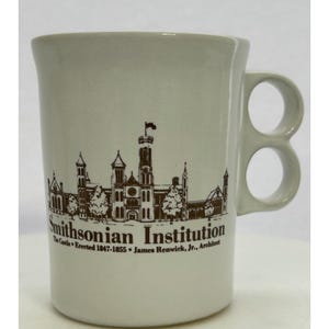 May include: A cream-colored ceramic mug featuring a brown illustration of the Smithsonian Institution building. The mug has a unique handle with two circular openings. Text on the mug reads "Smithsonian Institution" and "The Castle • Erected 1847-1855 • James Renwick, Jr., Architect."