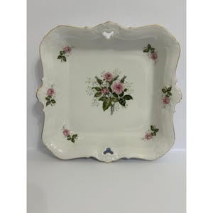 Pode incluir: Uma travessa quadrada de porcelana branca com borda dourada e um design floral de rosas. A travessa apresenta um buquê de rosas cor-de-rosa no centro e pequenos grupos de rosas nas bordas. As bordas têm um design decorativo ondulado.